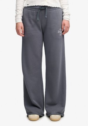 Personne portant un pantalon de survêtement gris ample avec une taille à cordon, un haut blanc et des chaussures beiges, debout avec les mains détendues de chaque côté.