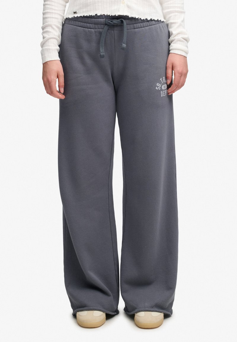 Personne portant un pantalon de survêtement gris ample avec une taille à cordon, un haut blanc et des chaussures beiges, debout avec les mains détendues de chaque côté.