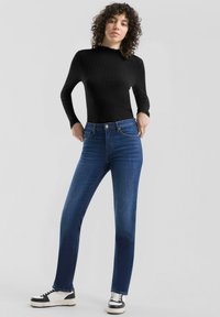 Zwarte geribbelde lange mouw top gecombineerd met blauwe rechte jeans, met vijf zakken en een neutraal gekleurde knop. Zwart-witte sneakers.