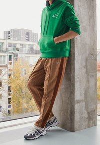 Felpa verde con tasca a marsupio, pantaloni sportivi marroni a fantasia con strisce laterali e sneakers bianche sportive con dettagli scuri.