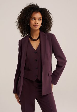 Donna con capelli ricci che indossa un completo viola scuro con un gilet abbottonato e una collana nera di perline, in posa con una mano in tasca.