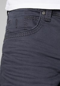 Gros plan sur la zone de la poche d'un jean en denim noir avec des coutures visibles, un bouton et une passants de ceinture, sur un fond de chemise blanche.