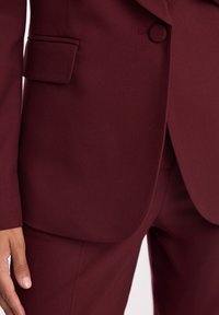 Bordowy blazer o teksturowanym wykończeniu, z pojedynczym zapięciem na guzik i kieszenią na piersi, w połączeniu z dopasowanymi spodniami w kant.