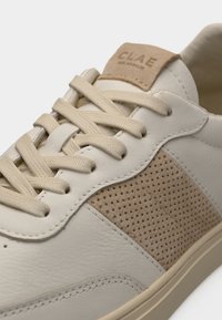 Κοντινό πλάνο μπεζ δερμάτινου αθλητικού παπουτσιού με διάτρητη πλευρική σουέτ επιφάνεια, κορδόνια στο ίδιο χρώμα και λογότυπο CLAE σε μπεζ ταμπέλα στη γλώσσα.