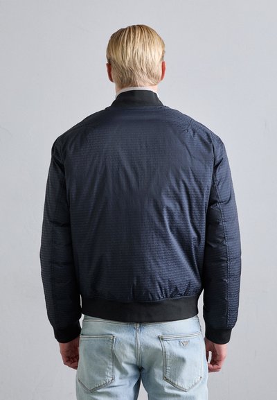Marinblå bombarjacka med ett texturerat mönster, ribbad mudd och nederkant, samt en slät yta, bärs över ljustvättade jeans.