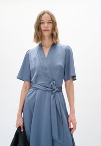 Hellblaue Wickelkleid mit kurzen Ärmeln, strukturiertem Stoff, tailliertem Gürtel und einem Knoten-Detail vorne, das eine fließende Silhouette betont.