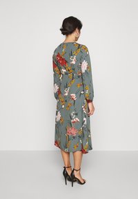 Mujer con cabello oscuro recogido en un moño, vestida con un vestido midi verde grisáceo con estampado floral y tacones altos negros con tiras en los tobillos, de pie y mirando hacia atrás.