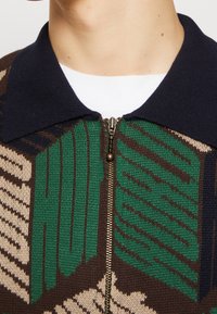 Stickad zip-up-tröja med en marinblå krage, prydd med ett geometriskt mönster i grönt, brunt och beige. Gulddragkedjedetalj.