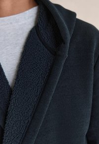 Tmavomodrá bunda s kapucňou s textúrovaným golierom z sherpy a rebrovaným materiálom, cez svetlošedé tričko.