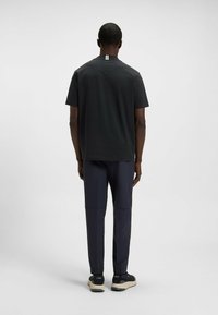 Man staat met zijn rug naar de camera, gekleed in een zwart T-shirt met witte strepen op de kraag en donkerblauwe joggingsbroek met zwarte sneakers.