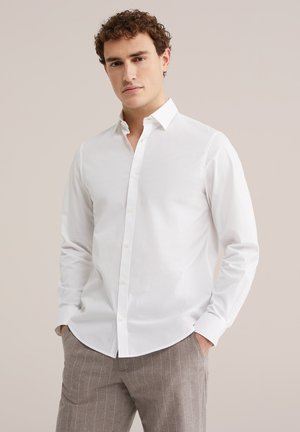 Muškarac s kovrčavom kosom u bijeloj košulji s gumbima i sivim hlačama na pruge, stoji s rukama u džepovima na neutralnoj pozadini.