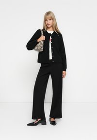 Gilet noir en tricot avec des boutons, t-shirt graphique blanc, pantalon noir ample et chaussures plates noires. Un sac texturé avec un motif serpent.