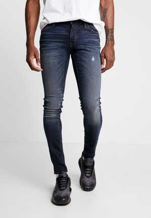 Homme portant un jean slim bleu marine avec des détails délavés, une chemise blanche, des baskets noires et des tatouages visibles sur les bras, sur un fond uni.