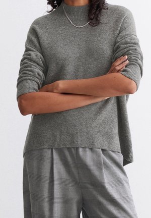Personne portant un pull en maille gris aux manches retroussées et un pantalon à carreaux gris, bras croisés, avec une chaîne argentée visible.