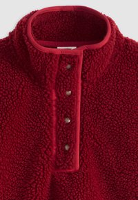 Camisola de lã polar vermelha com gola alta, apresentando uma superfície texturizada e quatro botões pressão na parte da frente. Bordas acabadas com uma cor contrastante.