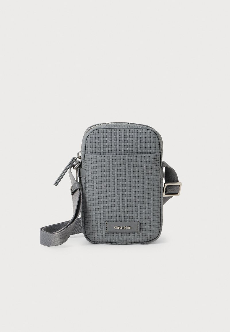 Pequena bolsa crossbody cinza com textura trançada, fecho de zip, alça ajustável e logotipo "Calvin Klein" no bolso frontal.