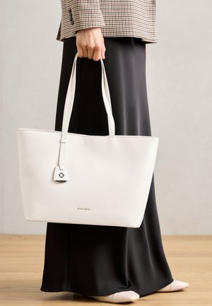 Sac fourre-tout blanc avec une texture lisse, de forme rectangulaire et des anses longues. Présente un petit accent en forme de serrure et le logo "ANNA FIELD" en bas.