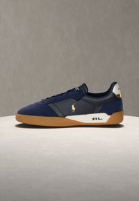 Zapatilla azul marino con suela de goma, lengüeta blanca en el talón, logo Polo dorado en el lateral y las iniciales "RL" en el panel blanco de la entresuela.