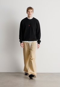 Μαύρο φούτερ με κόκκινο λογότυπο, λευκή μπλούζα με στρογγυλή λαιμόκοψη από κάτω, καφέ παντελόνι σε φαρδιά γραμμή και μαύρα παπούτσια, φορεμένο από μοντέλο που περπατά.