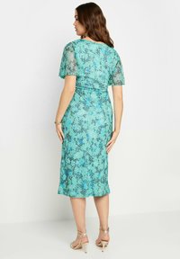 Robe turquoise avec des manches flottantes, arborant un motif floral en noir et bleu foncé. Elle arrive sous le genou, accompagnée de talons dorés clairs.