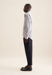 Camicia bianca con un piccolo motivo a pois neri, abbinata a pantaloni blu navy e scarpe eleganti nere, vista di lato.