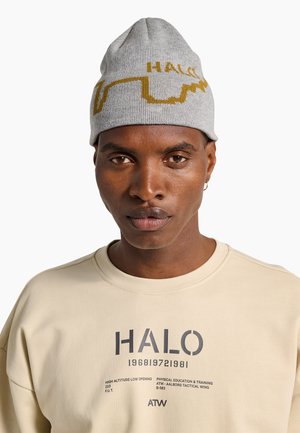 Homme portant un bonnet gris tricoté avec le texte jaune « HALO » et un sweat-shirt beige imprimé avec « HALO » et un texte de formation technique, faisant face à l'avant.