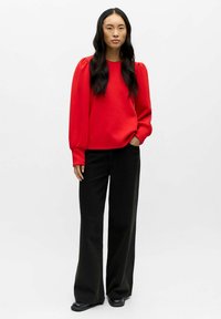 Roter Pullover mit langen Puffärmeln, kombiniert mit hochgeschnittenen schwarzen Schlaghosen und schwarzen Slippern, vor einem weißen Hintergrund stehend.