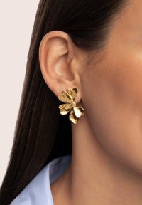 Orecchini floreali in oro con un design testurizzato, caratterizzati da forme di petali e da una perla centrale, indossati su un orecchio contro una pelle liscia.