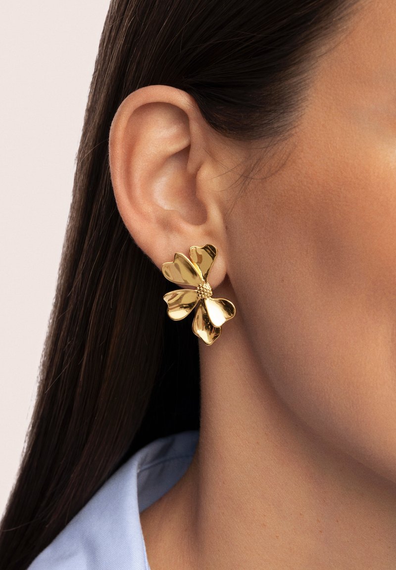 Orecchini floreali in oro con un design testurizzato, caratterizzati da forme di petali e da una perla centrale, indossati su un orecchio contro una pelle liscia.