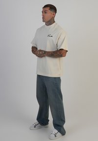 Beige oversized t-shirt med korte ærmer og høj halsudskæring. Udstyret med sort broderet logo. Båret med lyseblå jeans og grå sneakers.