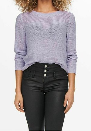 Femme portant un pull en maille violet clair rentré dans un pantalon noir taille haute avec quatre boutons visibles et des poches avant.