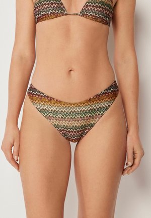 Kobieta ubrana w bikini w wielokolorowy wzór zygzaka, z trójkątnym topem i niskim stanem, na jednolitym jasnym tle.