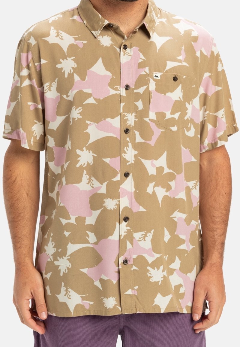 Homme portant une chemise à manches courtes à boutons avec des motifs floraux abstraits beiges, roses et blancs, et une poche poitrine.