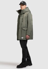 Oliven grøn polstret parka med høj krave, store forlommer og snoretræk. Bæret med sorte jeans og kraftige sorte støvler.