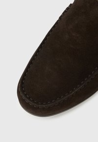 Σκούρο καφέ μοκασίνι από καστόρι με ορατή ραφή κατά μήκος της στρογγυλής μύτης και της άκρης, σε λευκό φόντο.