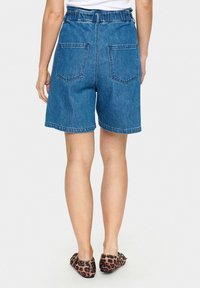 Vidējas zilas džinsa šorti ar elastīgu jostu, diviem aizmugures kabatām un taisnas kājas griezumu, kombinēti ar leoparda raksta apaviem.