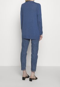 Modrý svetr s dlouhým rukávem, kulatým výstřihem, žebrovaným lemem a knoflíkovými detaily na rukávech, kombinovaný s modrými džíny s mid-rise pasem a černými sandály na blokovém podpatku.