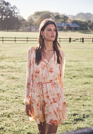 Robe florale en pêche clair avec des volants, des manches trois-quarts et une taille à cordon. Tissu doux et transparent avec un design fluide.
