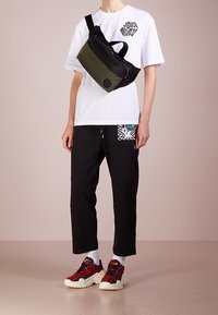 Borsa a tracolla nera e oliva con tracolla regolabile; indossata sopra una maglietta bianca e pantaloni joggers neri. Sneakers a quadri rossi con suole spesse.