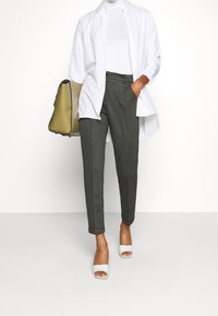 Grijs gemêleerde broek, witte getailleerde coltrui, oversized witte blouse met knopen, witte haksandalen en een grijs-groene handtas.