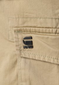 Beige tyg med en jämn textur, featuring en ficka och en svart metallsymbol med ordet "RAW." Vita stygn synliga längs kanterna.