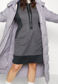 Femme portant une robe zippée grise associée à un long manteau matelassé lavande, debout les bras écartés sur fond blanc.