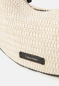 Struktūruotas beige audinio rankinės su juodu etiketė, kurioje užrašyta "Calvin Klein." Suapvalinta forma su tamsiomis akcentais palei kraštus.