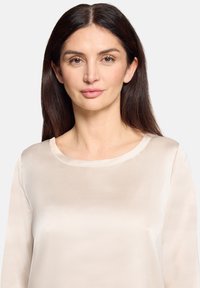 Hellbeiges Satinoberteil mit rundem Ausschnitt und langen Ärmeln, das eine glatte Textur und ein minimalistisch gestaltetes Design aufweist.