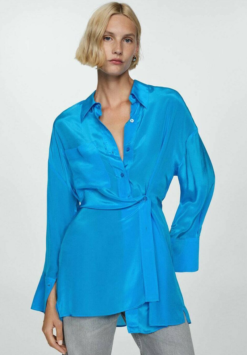 Blauwe satijnen blouse met een kraag, lange mouwen, knoopsluiting en een gestrikte taille. Bevat een zak aan de linkerkant en heeft een gladde textuur.
