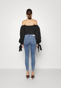 Top negro fuera de los hombros con mangas voluminosas y detalles de lazo, combinado con jeans ajustados azules de cintura alta y zapatos de tacón negros.