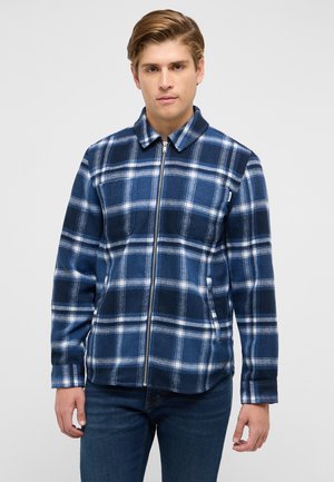 Junger Mann, der eine blau-weiß karierte Reißverschlussjacke und dunkle Jeans trägt, vor einem einfarbigen hellgrauen Hintergrund steht.