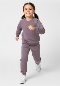 Jasnofioletowa bluza i pasujące spodnie dresowe wykonane z miękkiego materiału, z prążkowanymi mankietami i luźnym krojem. Białe sneakersy dopełniają strój.