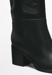 Zwarte leren enkelboot met een strak ontwerp, voorzien van een stevige blokhak en een soepele textuur, met gestikt detail langs de onderrand.