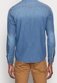 Camicia in denim di colore azzurro chiaro con maniche lunghe, polsini abbottonati, orlo dritto e dettagli sul giromanica posteriore. Abbinata a pantaloni kaki.